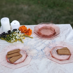 Può includere: Quattro piatti ottagonali di vetro rosa su una tovaglia bianca. Due piatti hanno biscotti. Due candele bianche e un mazzo di fiori sono sullo sfondo.