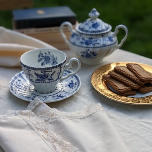 Puede incluir: Un juego de té vintage con diseños florales azules, que incluye una taza, un platillo y una azucarera. Un plato con borde dorado contiene varias galletas rectangulares. Una servilleta de lino color crema con ribete de encaje está en primer plano.