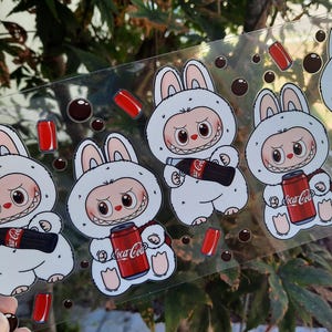May include: A clear sticker sheet featuring cartoon bunnies in white hooded outfits, each holding a Coca-Cola bottle or can. The bunnies have red eyes and a mischievous grin. Red cola cans and brown bubbles are scattered around.