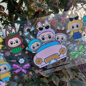 May include: A clear sticker sheet featuring cartoon characters in bunny costumes, driving a pink car. The sheet includes colorful bows, rainbows, cupcakes, and candies. The characters have large eyes and are wearing sunglasses.