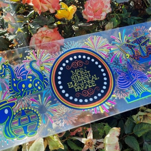 May include: A clear, rectangular sticker featuring colorful illustrations and the text "Main Street Electrical Parade." The design includes a dragon, turtle, and other whimsical figures, all set against a backdrop of fireworks.