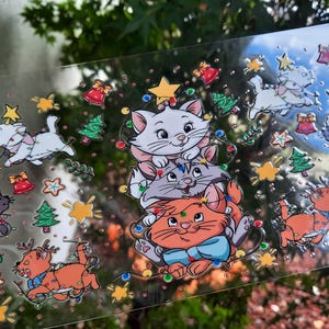 May include: Clear sticker sheet featuring cartoon cats, Christmas trees, bells, and stars. The cats are in various poses, some wearing Santa hats and wrapped in Christmas lights. The sticker sheet has a festive, holiday theme.