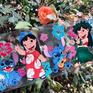 May include: A clear sticker sheet featuring cartoon characters Lilo and Stitch. The sheet includes images of Lilo in a red dress, Stitch playing a ukulele, and other characters with floral accents in pink and blue. The background shows green foliage and a rose.