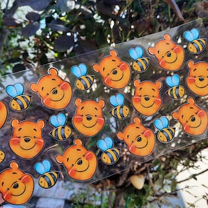 Peut inclure: Une feuille d'autocollants transparents avec des images de dessins animés d'un ours orange souriant et d'abeilles jaunes et noires avec des ailes bleues. Les autocollants sont disposés en motifs répétés.