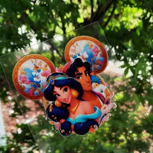 May include: A clear sticker featuring a colorful illustration of Princess Jasmine and Aladdin from Disney's Aladdin. The design incorporates Mickey Mouse ears with floral and ornate details. The characters are depicted with vibrant colors and detailed features.