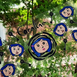 May include: A clear, rectangular sticker featuring the name "Coraline" in black script. The sticker includes illustrations of the character Coraline with blue hair and black keys, set against a blurred green background.
