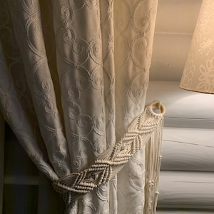 May include: White curtains with a delicate floral pattern are tied back with a macrame curtain tie. The tie is made of natural cotton rope and has a wooden ring at the top.