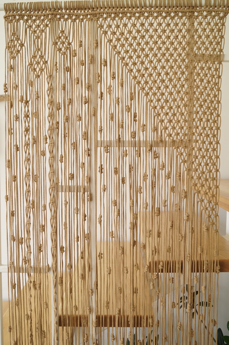 May include: A macrame wall hanging made of natural beige colored cord. The hanging features a geometric pattern with a diamond shape in the center.