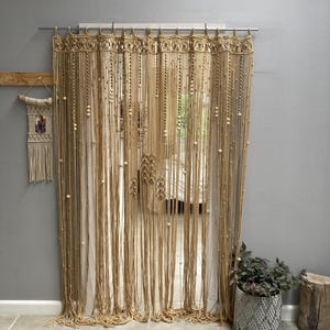 May include: A macrame curtain with wooden beads hangs in a doorway. The curtain is made of natural fiber rope and has a woven pattern. The curtain is beige in color and is hanging from a metal rod.