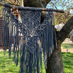May include: A grey macrame wall hanging with a geometric pattern and fringe. The hanging is made of cotton cord and is attached to a branch.