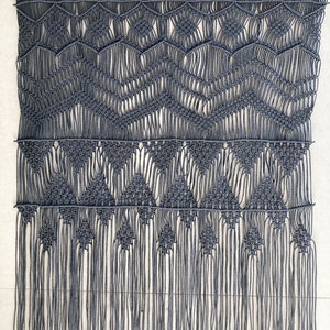 Pode incluir: Uma tapeçaria de macramé cinza com um padrão geométrico e franjas. A tapeçaria é feita de cordão de algodão grosso e tem um design tecido.