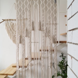 Pode incluir: Uma tapeçaria de macramé branca com um padrão geométrico e franjas. A tapeçaria é feita de cordão de algodão e está pendurada em uma barra de madeira.