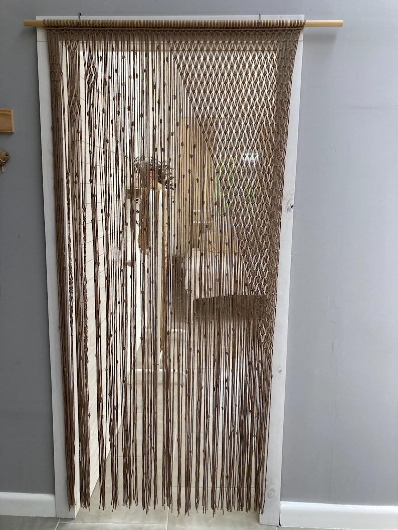 May include: A brown macrame curtain with wooden beads hangs in a doorway. The curtain is made of knotted rope and has a diamond pattern.