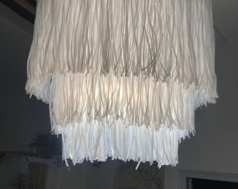 May include: A white, multi-tiered lampshade with a fringe design. The lampshade is made of thin, white fabric strips that hang down in a layered pattern.
