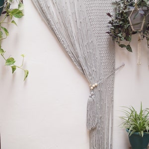 May include: A gray macrame wall hanging with wooden beads and tassels. The hanging is made of thick cotton cord and is hung from a wooden dowel. The hanging is in front of a white wall with plants on either side.