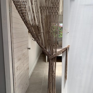 Puede incluir: Una cortina de macramé marrón cuelga en una puerta, con una puerta de madera blanca a la izquierda y una pared blanca a la derecha. La cortina está hecha de cuerdas anudadas y tiene un patrón geométrico.