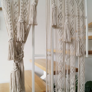 May include: Cream-colored macrame curtain with intricate knotting and tassel details. The curtain hangs in front of a staircase with wooden steps and a white metal railing. The macrame design adds a bohemian touch to the interior.