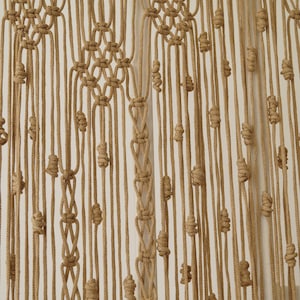 May include: A macrame wall hanging made with natural brown cord. The cord is knotted in a pattern that creates a series of small, round shapes. The hanging is a light brown color and has a natural, rustic look.
