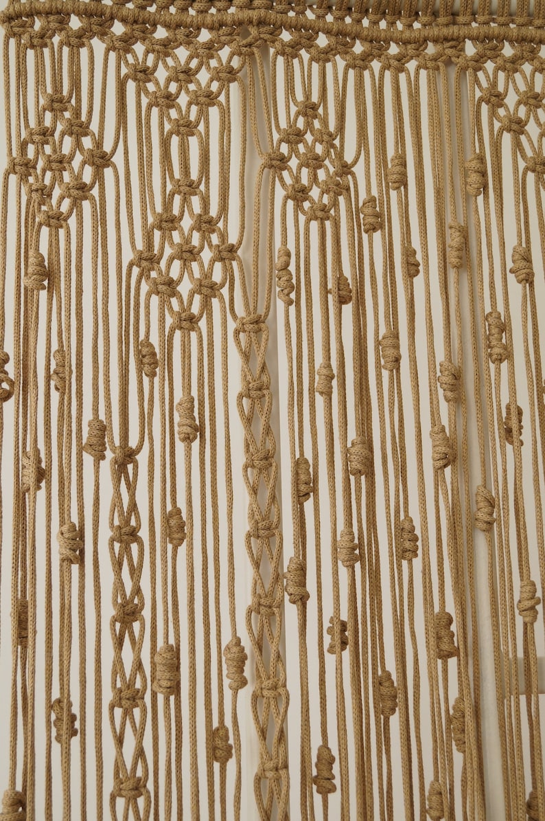 May include: A macrame wall hanging made with natural brown cord. The cord is knotted in a pattern that creates a series of small, round shapes. The hanging is a light brown color and has a natural, rustic look.