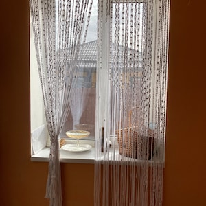 May include: White macrame curtain hanging in a window with a wicker basket and a glass dish on a white windowsill. The window is behind the curtain and shows a view of a building with a tiled roof.