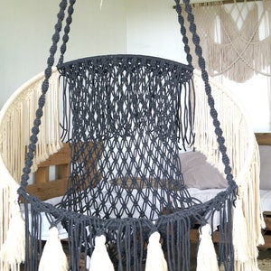 May include: A black macrame hanging chair with white tassels. The chair is suspended from the ceiling by two thick ropes.