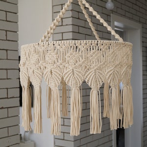 May include: A white macrame hanging lamp with a circular shape and fringe tassels. The lamp is hanging from a white ceiling in a room with white brick walls.