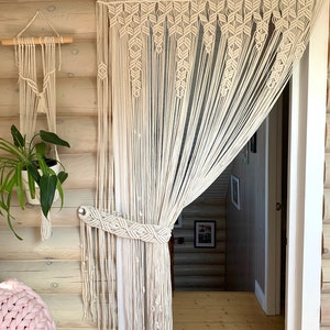 May include: A white macrame curtain hangs in a doorway, with a smaller macrame wall hanging on the left side of the image. The curtain is made of knotted cords and has a decorative pattern at the top.
