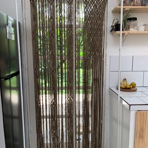 Pode incluir: Uma cortina de macramé marrom com um padrão geométrico pendurada em uma porta. A cortina é feita de corda grossa e tem uma franja na parte inferior.