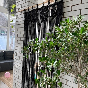 Peut inclure: Une tenture murale en macram&eacute; noire avec un motif g&eacute;om&eacute;trique. La tenture est faite d'un cordon noir &eacute;pais et est suspendue &agrave; une tringle en bois. La tenture est devant un mur en briques blanches.