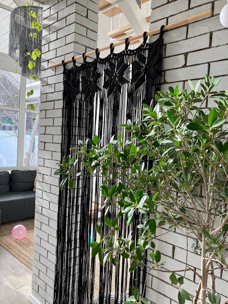 Peut inclure: Une tenture murale en macram&eacute; noire avec un motif g&eacute;om&eacute;trique. La tenture est faite d'un cordon noir &eacute;pais et est suspendue &agrave; une tringle en bois. La tenture est devant un mur en briques blanches.