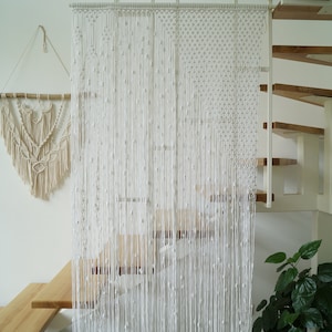 May include: A white macrame curtain hangs in front of a wooden staircase. The curtain is made of a series of knotted cords that create a delicate and airy look.