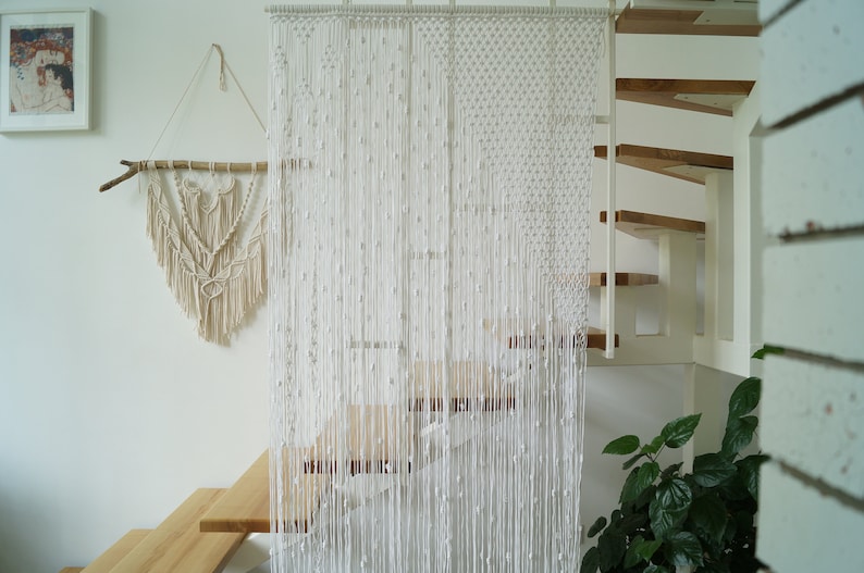 May include: A white macrame curtain hangs in front of a wooden staircase. The curtain is made of a series of knotted cords that create a delicate and airy look.