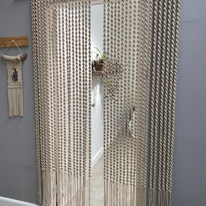 May include: Beige macrame door curtain with a fringe. The curtain, made of knotted rope, hangs in front of a doorway. A small macrame wall hanging is on the left. The door is open, revealing a room with a plant.