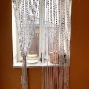 Puede incluir: Cortina de macramé blanca colgada en una ventana. La cortina está hecha de una serie de cordones anudados que cuelgan de una barra. La cortina está parcialmente abierta, revelando una vista de un edificio fuera de la ventana.
