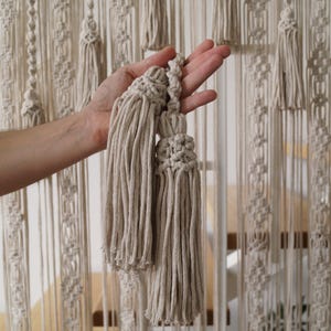 Pode incluir: Dois pingentes de macramé bege com uma parte superior de nó, segurados na mão contra um fundo de uma tapeçaria de macramé bege.