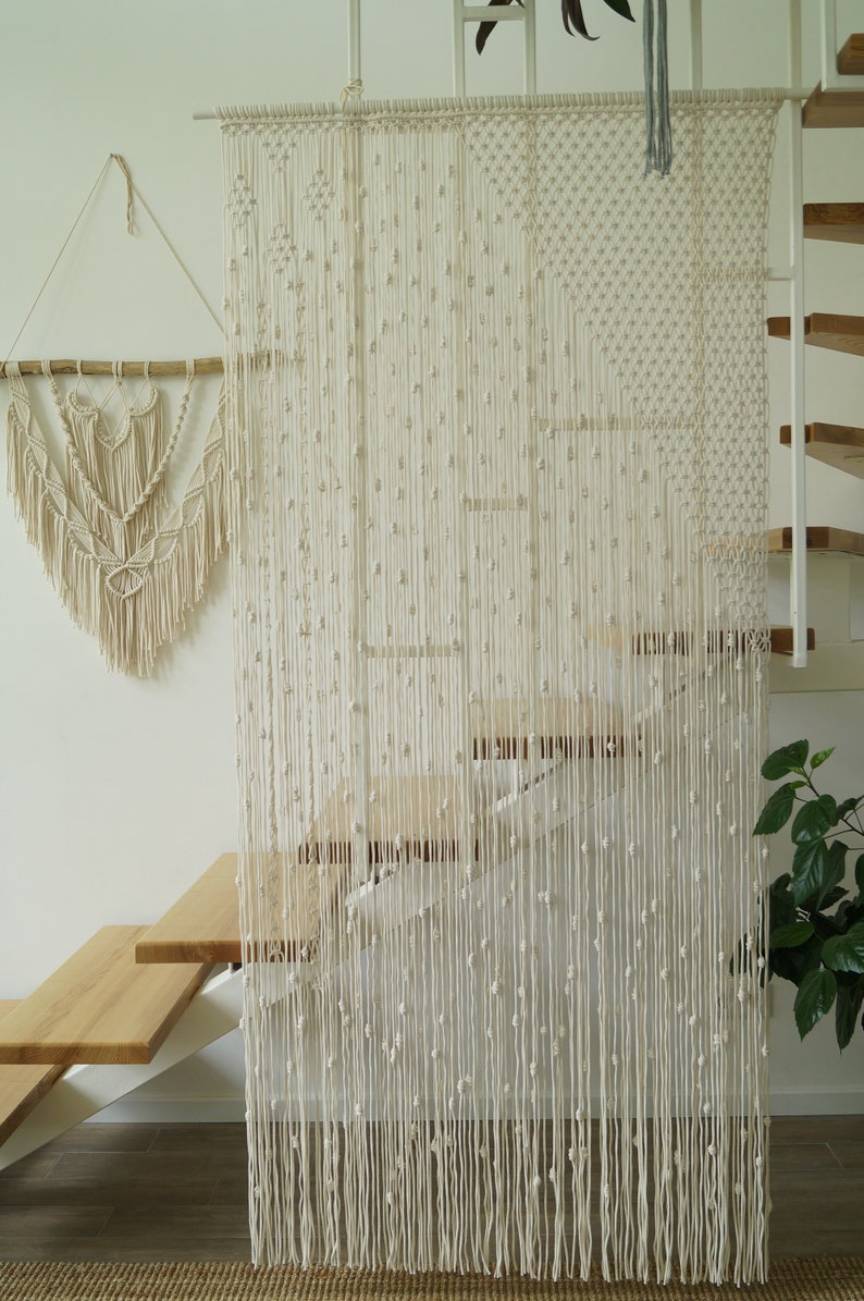 May include: A white macrame curtain with knotted fringe hangs in front of a wooden staircase. The curtain is made of natural fibers and has a bohemian style.