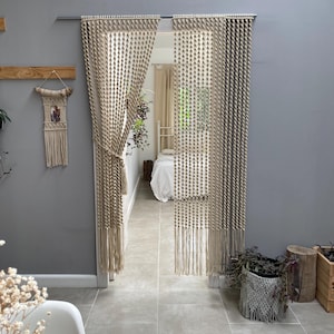 May include: Beige macrame curtains hanging in a doorway. The curtains feature a geometric design and fringe. A macrame wall hanging is on the left. A bedroom is visible through the doorway.