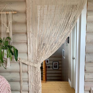 Op de afbeelding: Een crèmekleurig macramé gordijn met een houten stok aan de bovenkant. Het gordijn is gemaakt van geknoopte koorden en heeft een parelmoerontwerp. Het gordijn hangt in een deuropening.