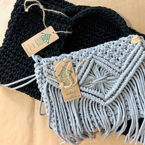 Peut inclure: Deux sacs en crochet, un noir et un gris avec des franges. Le sac gris a un motif géométrique et les deux sacs ont des étiquettes avec le texte "Your Knitted Home" et un logo.