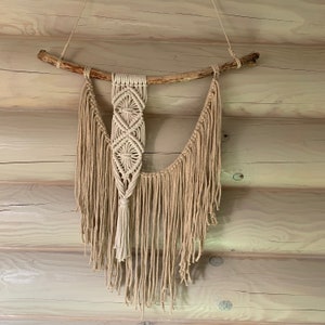 May include: A macrame wall hanging featuring a natural wood branch and beige cotton fringe. The macrame design is geometric with a tassel at the bottom.