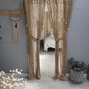 Pode incluir: Um par de cortinas de macramé bege com contas de madeira penduradas em uma porta. As cortinas são feitas de corda grossa e têm uma franja na parte superior.