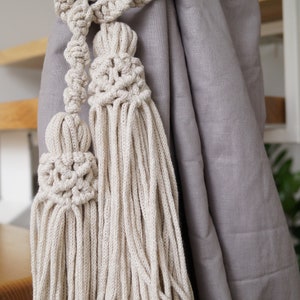 Pode incluir: Um par de amarrações de cortina de macramé bege com borlas. As amarrações estão amarradas em torno de uma cortina cinza.