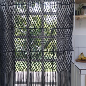 Puede incluir: Cortina de macramé negra colgada frente a una ventana. La cortina presenta un tejido con patrón de diamantes con flecos en la parte inferior. La ventana revela una vista del follaje verde.
