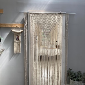 May include: A white macrame curtain with wooden beads hangs from a metal rod in a doorway. The curtain is made of knotted rope and has a geometric pattern. The curtain is hanging in front of a white door and a white tiled floor.