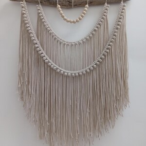 May include: A beige macrame wall hanging with a driftwood branch, two curved rows of knotted rope, and long fringe. A string of wooden beads hangs below the branch. This handcrafted piece adds a bohemian touch to any space.