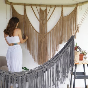 Pode incluir: Uma mulher está em frente a uma tapeçaria de macramê de parede com uma rede cinzenta em primeiro plano. A tapeçaria de parede é feita de corda de fibra natural e tem um design de franjas. A rede também é feita de corda e tem um design de franjas.