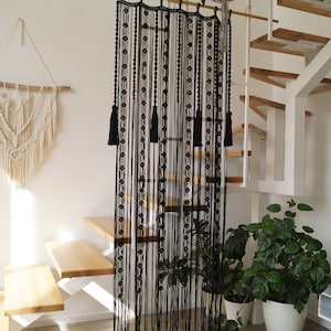 May include: Black macrame wall hanging with tassels, hanging from a wooden dowel rod. The wall hanging is in front of a white wall and a wooden staircase.
