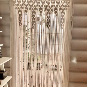 Pode incluir: Uma cortina de macramé branca com um padrão geométrico pendurada em uma porta. A cortina é feita de cordão de algodão grosso e tem uma franja na parte inferior.