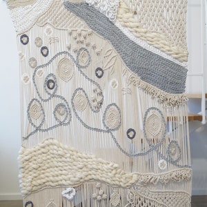 May include: A large macrame wall hanging with a variety of textures and patterns. The hanging is made with natural coloured cord and features a grey and white geometric design.