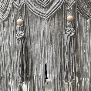 May include: Gray macrame wall hanging with two tassels and wooden beads. The macrame is made with a thick cord and has a geometric pattern.